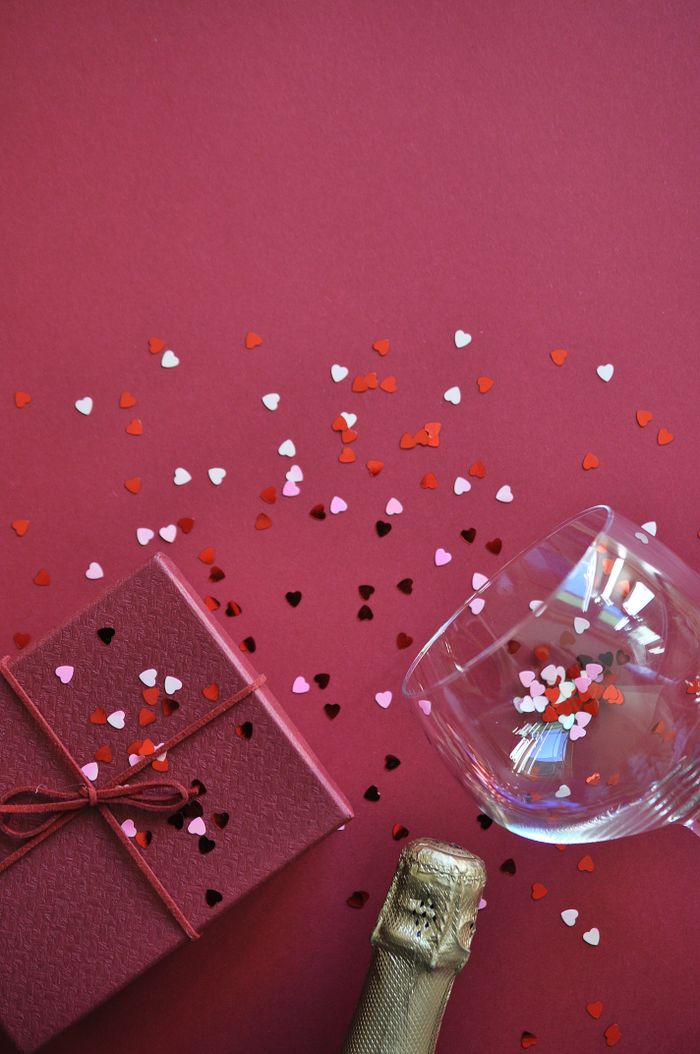 Romantic setting with champagne, gift box, and heart confetti on red background.